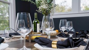 A dining table set with wine glasses, plates, and cutlery, with a vase of flowers and a bottle of wine in the background. - Lockeland Luxe｜Walk to 5 Points｜2 mi to Broadway (Nashville)