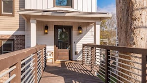 A wooden deck leading to a white building with a dark entrance door, two wall-mounted lanterns, and a brick wall adjacent to the building. - Lockeland Luxe｜Walk to 5 Points｜2 mi to Broadway (Nashville)