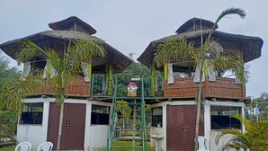 Een gebouw met twee verdiepingen, elk voorzien van een rieten dak en een balkon. Een centrale trap leidt naar de bovenverdieping.