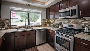 A modern kitchen with dark wood cabinets, stainless steel appliances, and a large window with an American flag. - Hidden Gem - Spacious and Gorgeous Getaway by the Pool  (Palm Springs)