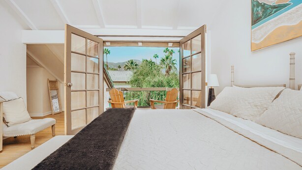 A bedroom with a large bed, a view of palm trees and mountains through open doors, a small balcony with chairs, and a painting on the wall. - Hidden Gem - Spacious and Gorgeous Getaway by the Pool  (Palm Springs)