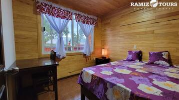 A wooden room with a bed, a nightstand, a desk, and a window with curtains.