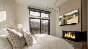 Un dormitorio moderno con una cama grande, chimenea, televisor montado en la pared y balcón con vistas a la montaña.