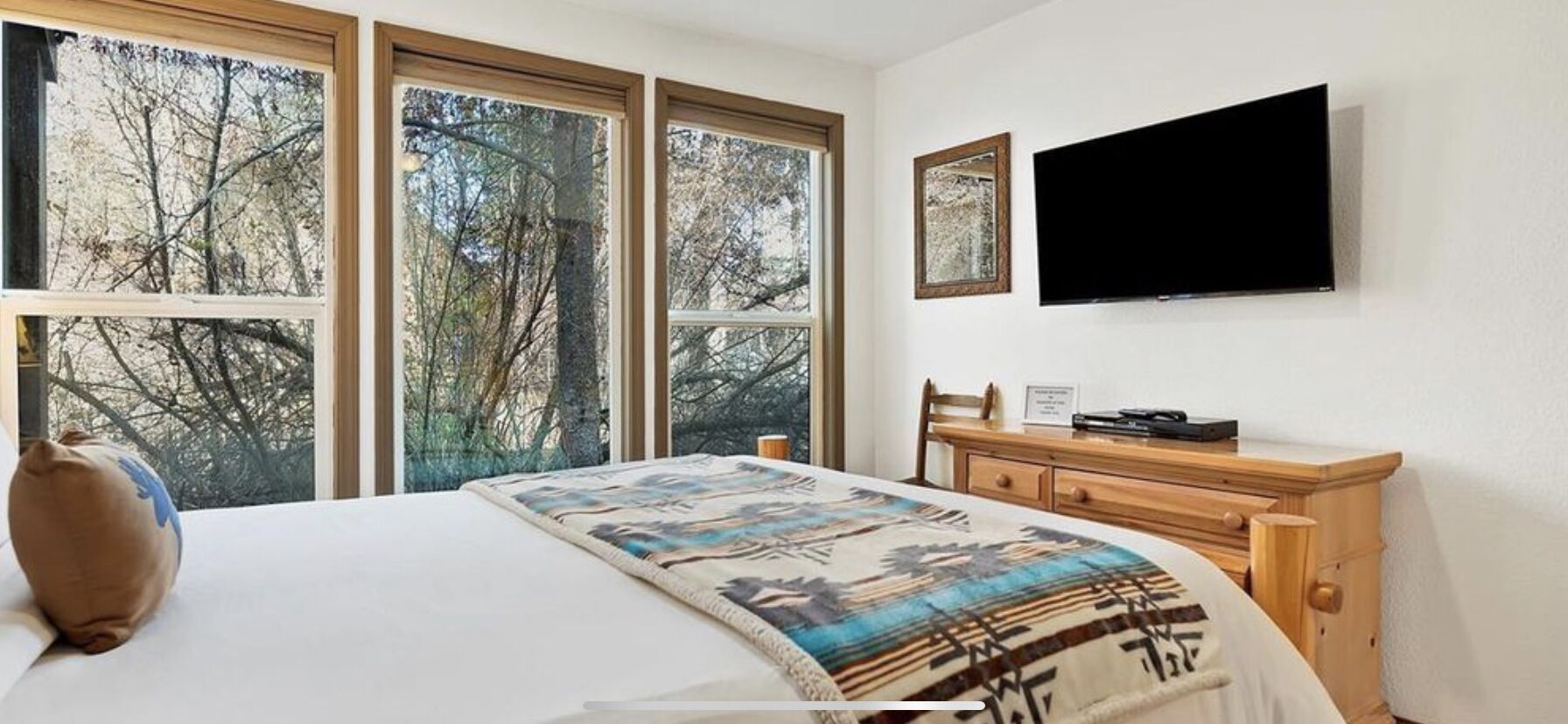 A bedroom with a bed, a dresser, a television, and large windows with a view of trees.