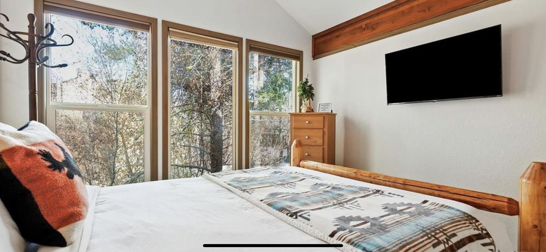 Une chambre avec une grande fenêtre, un lit recouvert d’une couverture à motifs, une commode en bois et un téléviseur à écran plat fixé au mur.