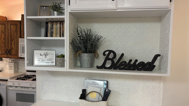Una cocina con armarios blancos, un cartel que dice 'Blessed', una planta en maceta y una cesta con una tarjeta y controles remotos.