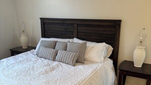 Una cama impecable con una colcha blanca texturizada y varios almohadones, con mesitas de noche de madera oscura y jarrones decorativos a cada lado.