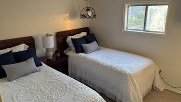 Une chambre avec deux lits, une table de chevet avec une lampe, un luminaire décoratif fixé au mur et une fenêtre donnant sur un paysage verdoyant.