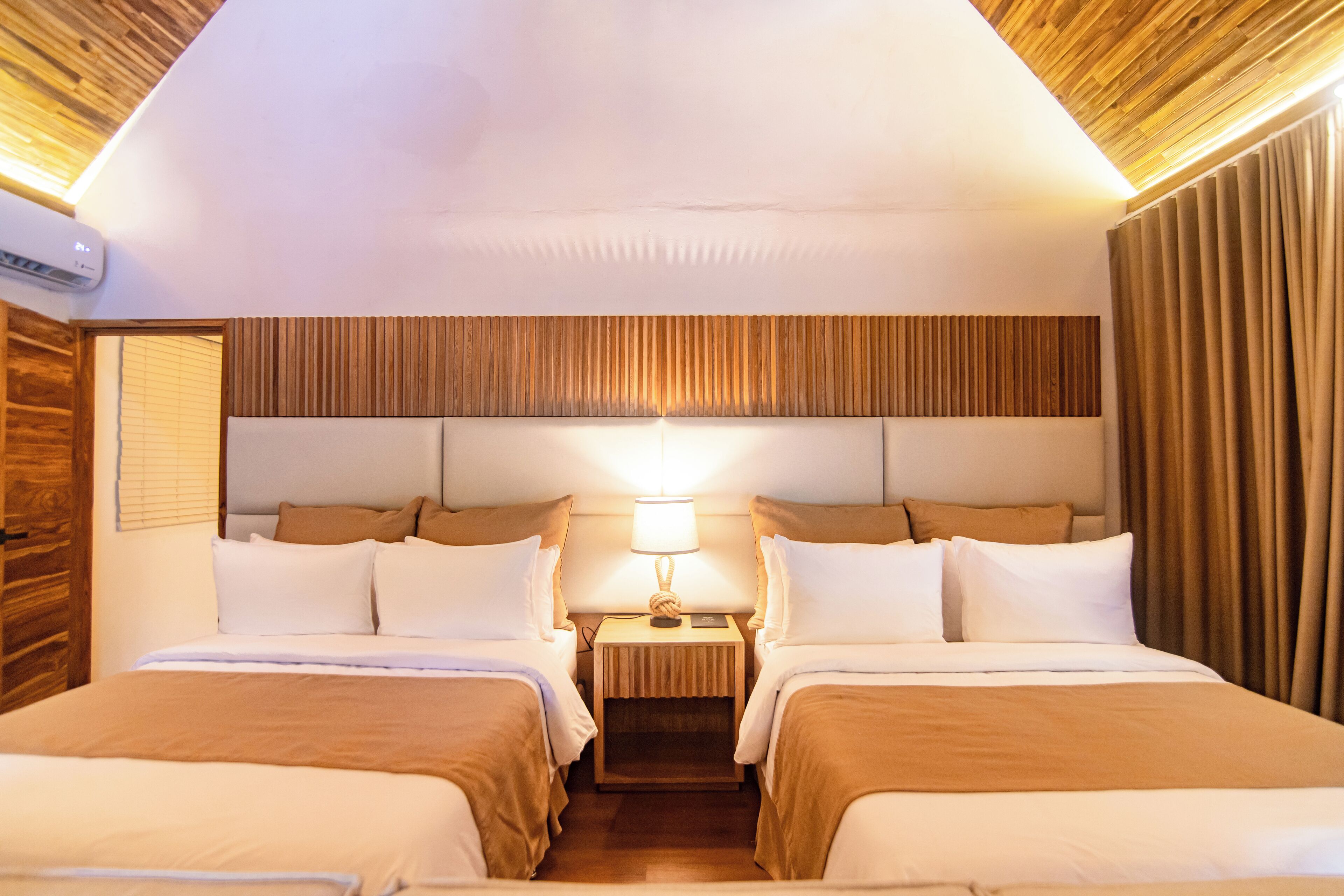 A hotel room with two beds, a wooden headboard, a bedside table with a lamp, and a window with blinds.