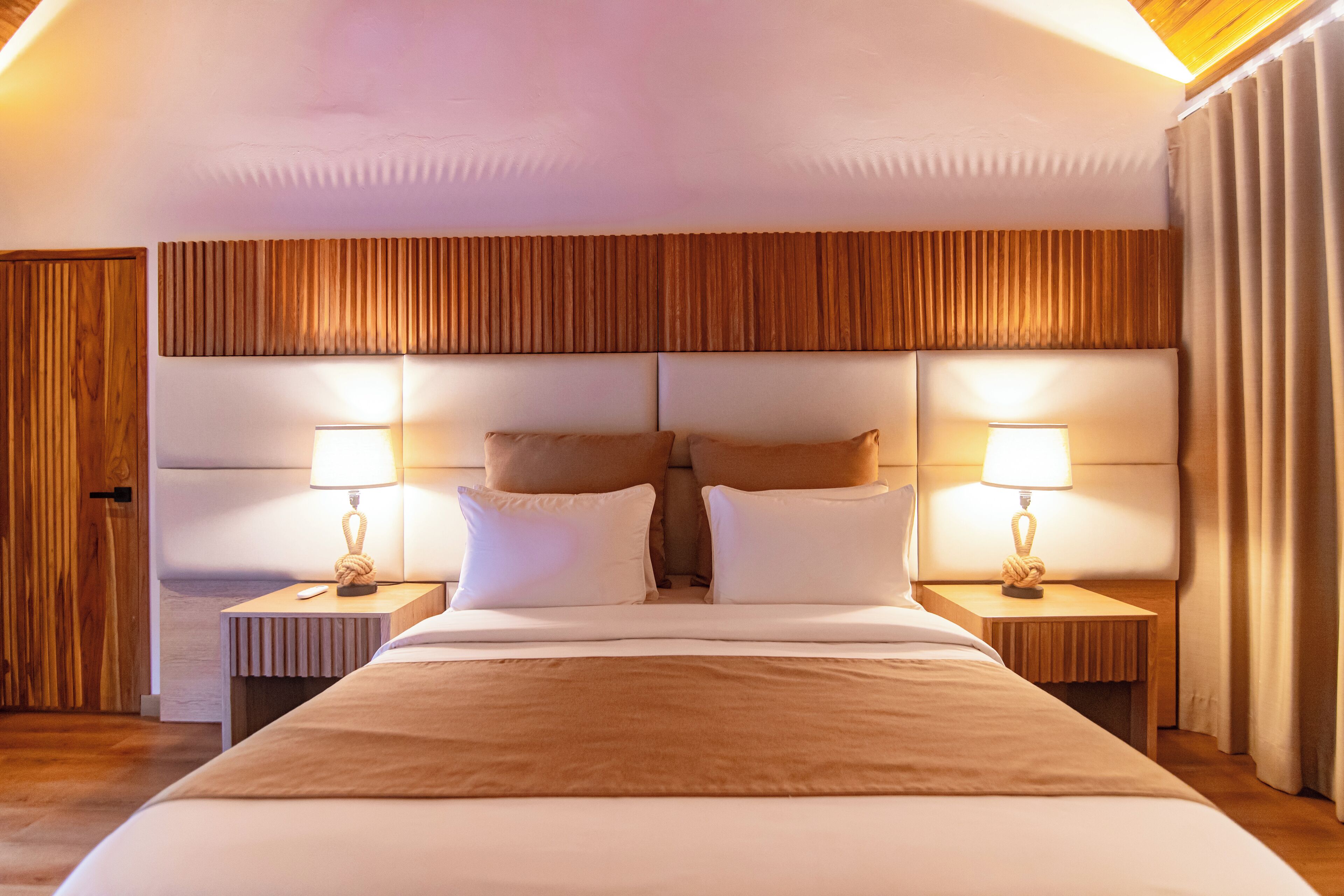 Una camera d'albergo con un letto grande, due comodini con lampade, una testiera in legno e una porta di legno.