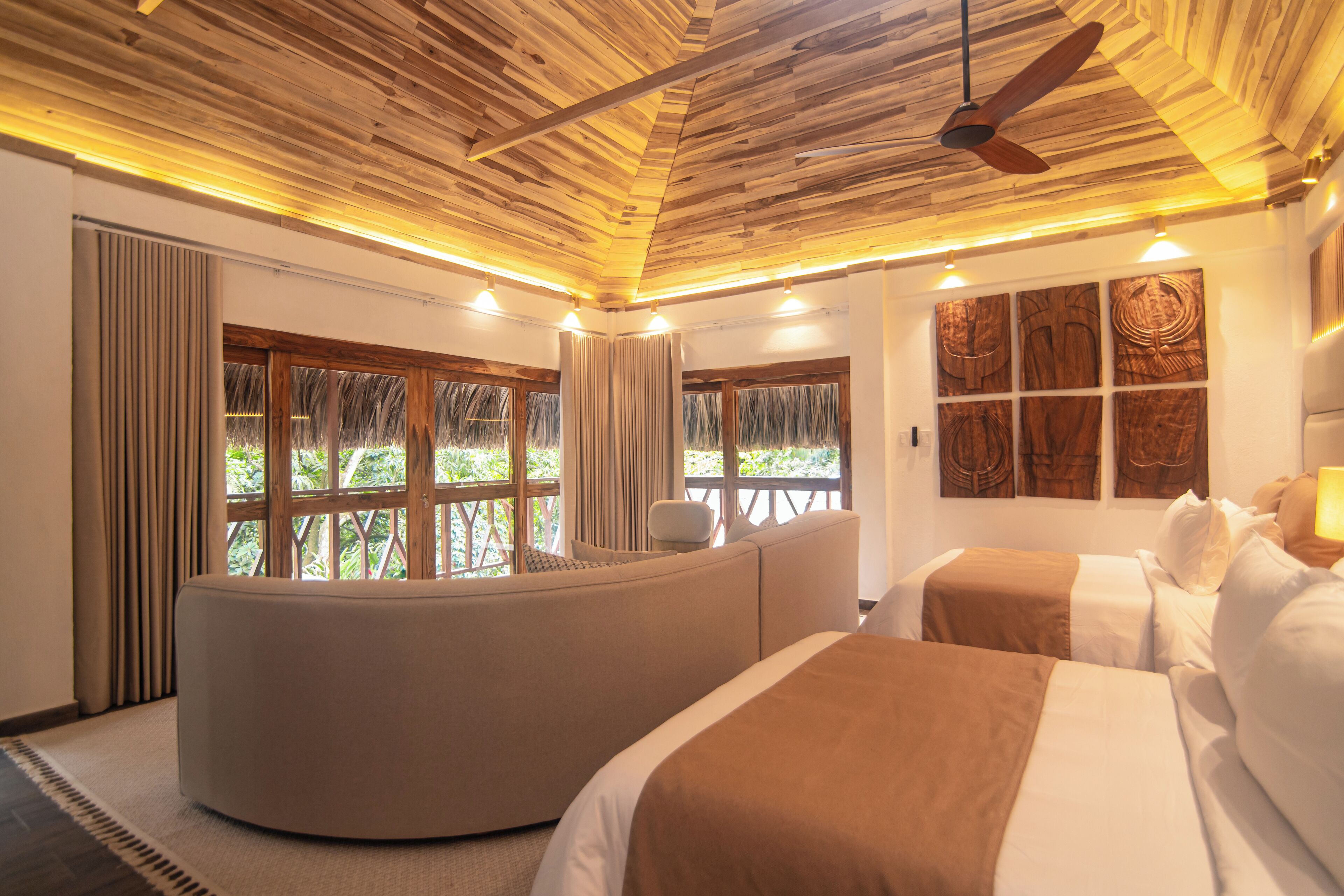 A hotel room with a curved seating area, two beds, a ceiling fan, and large windows with curtains.