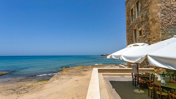 A seaside restaurant with outdoor seating, a stone building, and a view of the ocean.