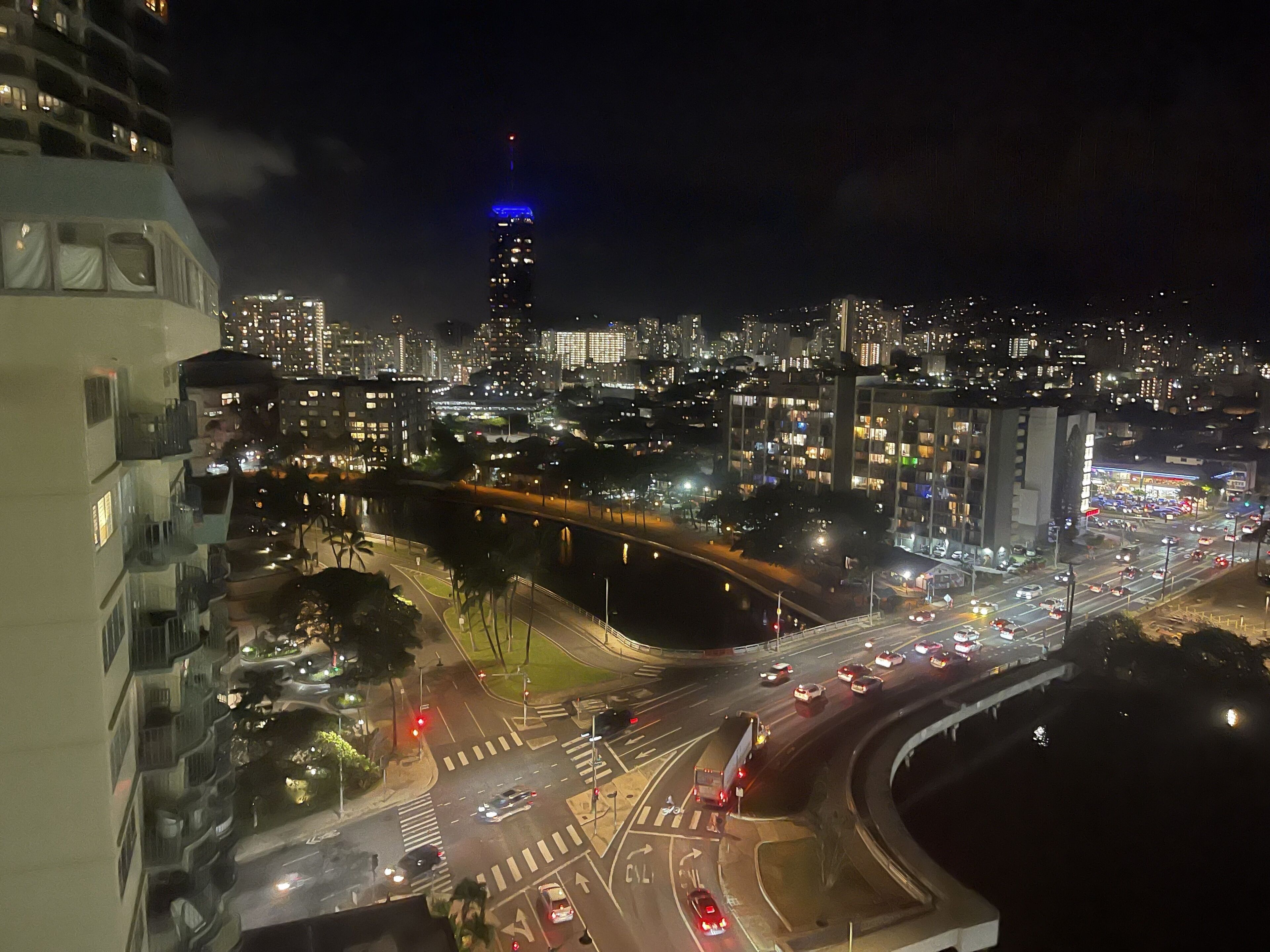 A nighttime cityscape with a prominent building with blue lighting on top, a busy intersection with multiple lanes, and a river running through the city.