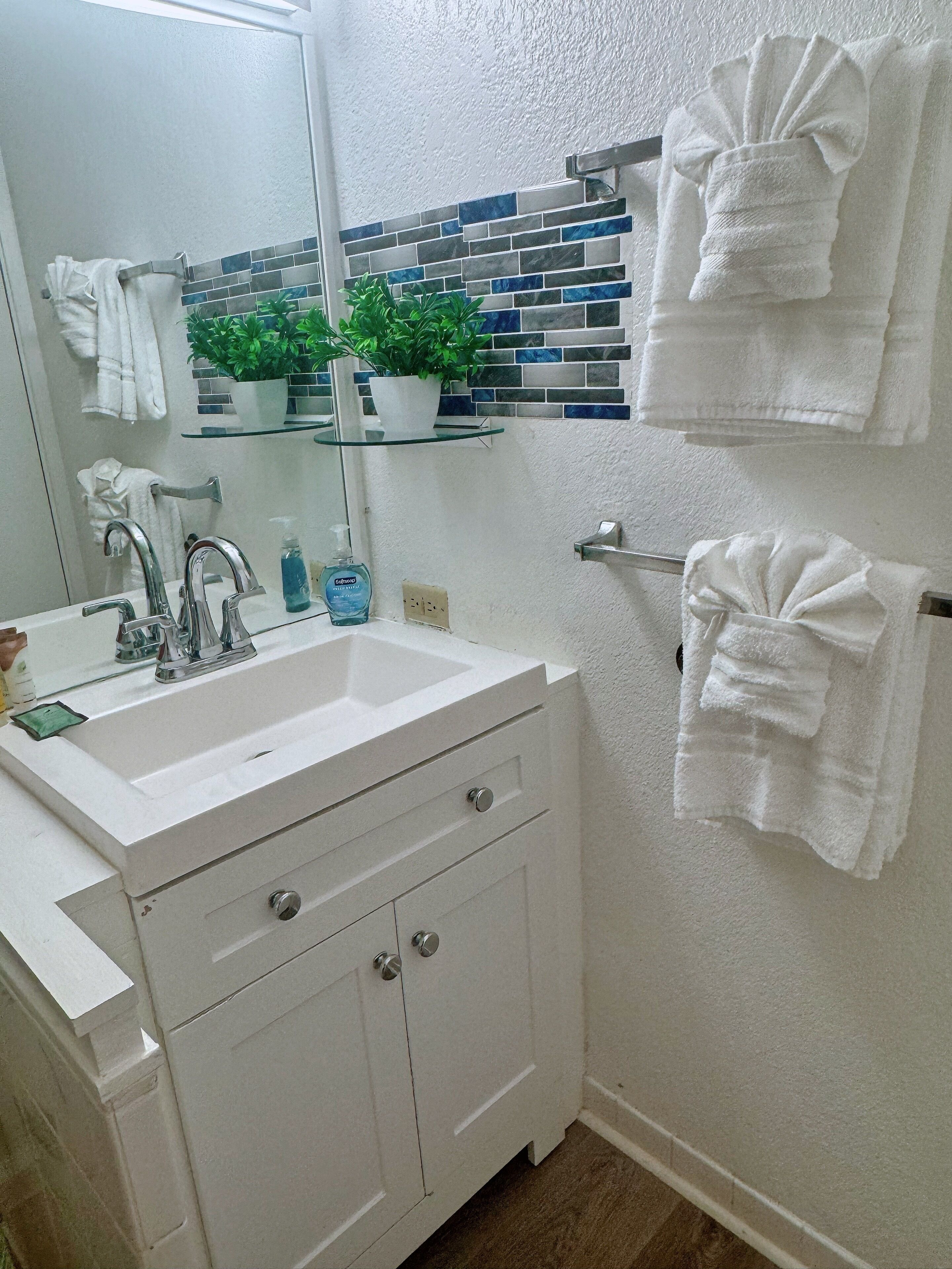 Une salle de bain avec un lavabo blanc, deux porte-serviettes, un miroir et une plante en pot posée sur une étagère.