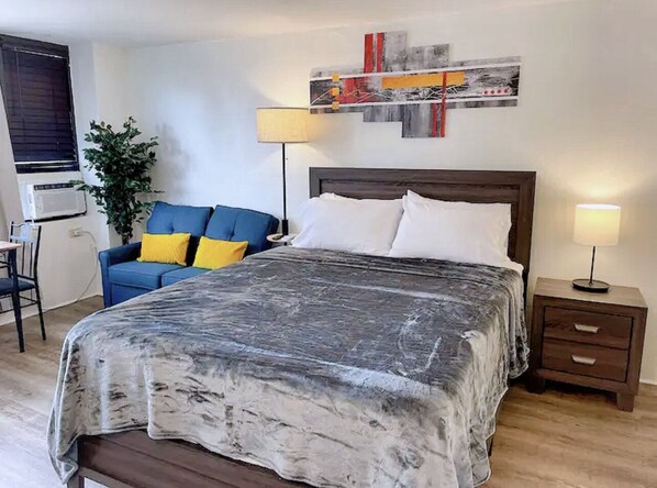 A bedroom with a large bed, a desk, a chair, a sofa with cushions, a lamp, and a wall art piece. - Cozy condo at Hawaiian monarch- no resort fee, free parking, near beaches, shop (Honolulu)