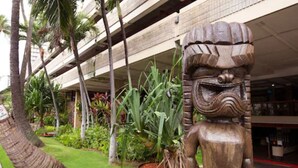 A statue of a figure with a beard and a headdress, surrounded by tropical plants and palm trees, in front of a multi-story building with a covered walkway. - Cozy condo at Hawaiian monarch- no resort fee, free parking, near beaches, shop (Honolulu)