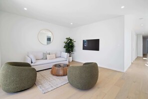 Ein modernes Wohnzimmer mit einem weißen Sofa, zwei grünen Poufs, einem runden Couchtisch, einem großen Spiegel, einer Topfpflanze und einem an der Wand montierten Flachbildfernseher.