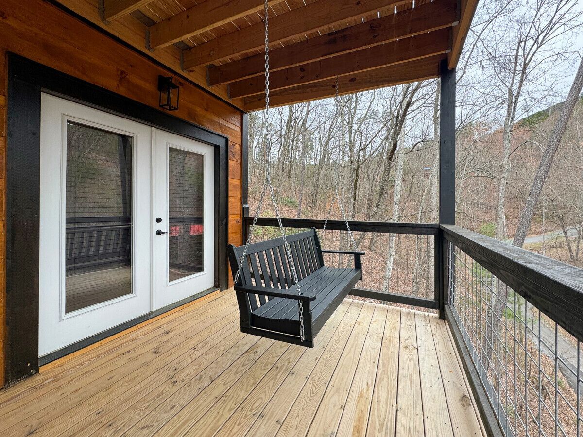 A wooden deck with a black swing, a glass door, and a view of trees.