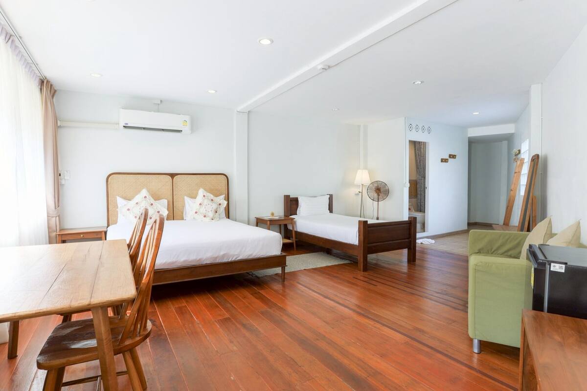 A hotel room with a wooden floor, a bed with a headboard, a bedside table, a lamp, a green sofa, and a small table with chairs.