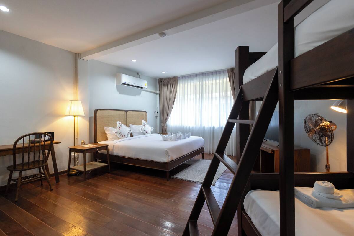 A hotel room with a bunk bed, a desk with a lamp, a bedside table, a chair, and a window with curtains.