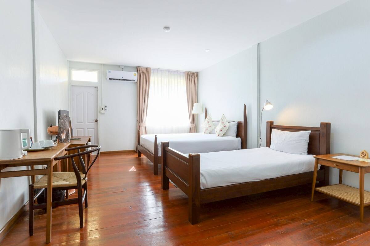 A hotel room with two beds, a desk, chairs, a lamp, and an air conditioning unit.