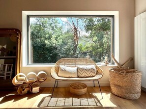 Una habitación con un ventanal grande, una silla de mimbre con cojines, una cesta tejida y una pequeña mesa de madera.