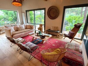 Een ruime woonkamer met een bank, stoelen, een salontafel en een groot raam met uitzicht op groen.