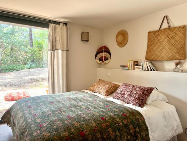 Een slaapkamer met een bed, kussens, een wandlamp, een geweven mand en uitzicht op groen buiten.