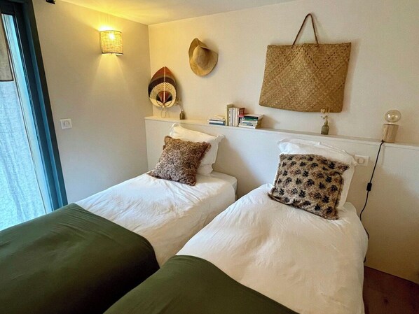 Een hotelkamer met twee enkele bedden, een wandlamp, een geweven mand en een kleine tafel met boeken en een vaas.