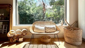 Un rincón acogedor para leer, con una silla de mimbre, una pequeña mesa redonda y vistas a un bosque verde exuberante a través de un ventanal grande.
