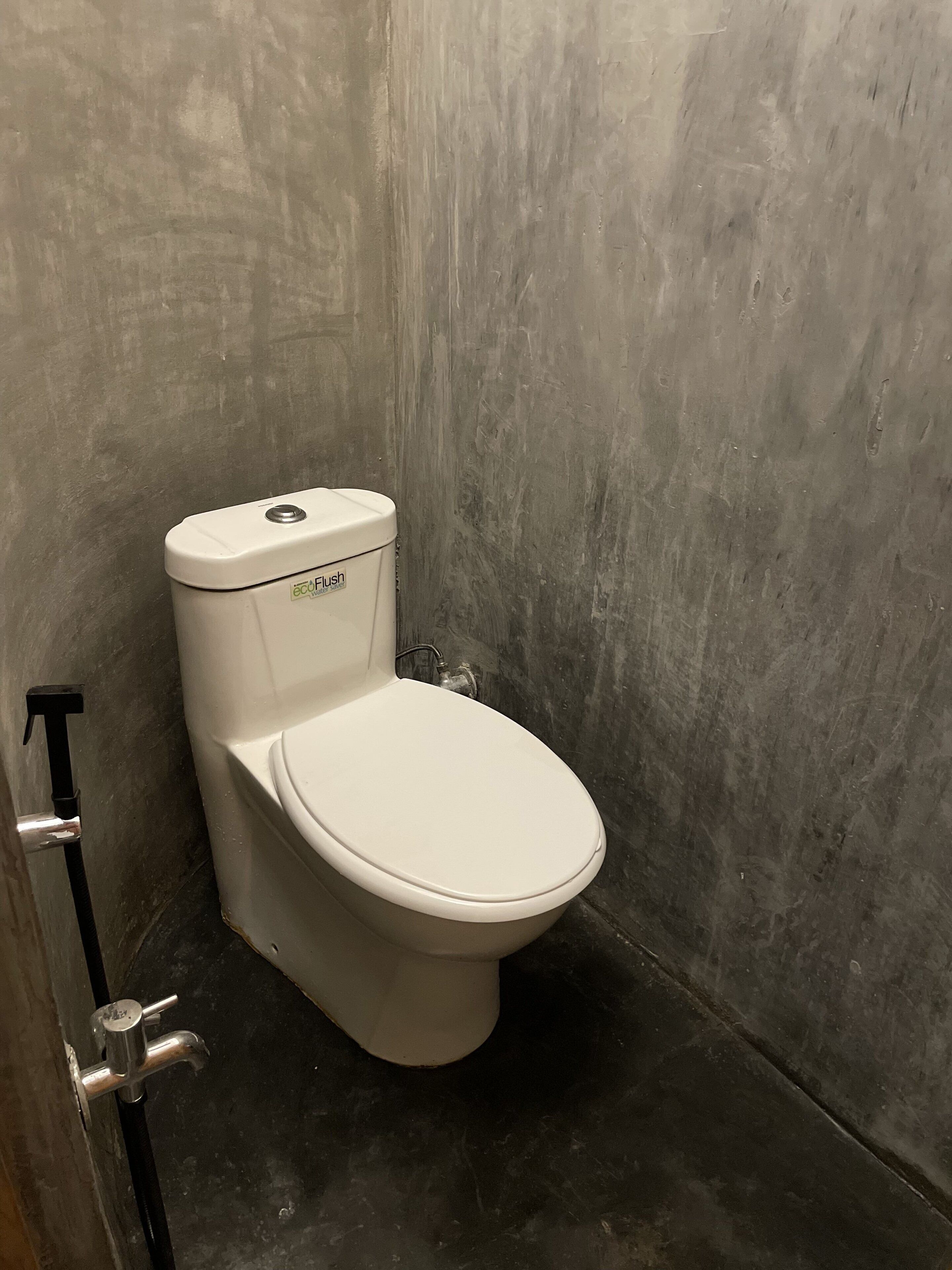 Un inodoro en un baño con pared de concreto y tubería metálica.
