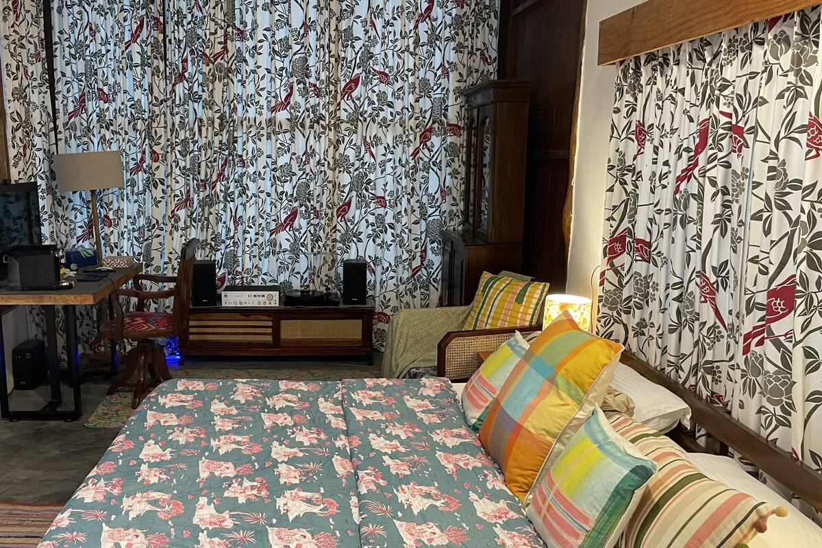 Un dormitorio con cortinas de estampado floral, una cama con almohadas coloridas, un escritorio de madera con una lámpara y una radio.
