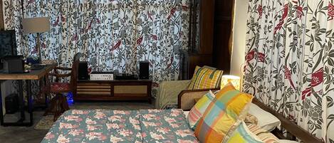 Une chambre à coucher avec des rideaux à motifs floraux, un lit agrémenté de coussins colorés, un bureau en bois avec une lampe et une radio.