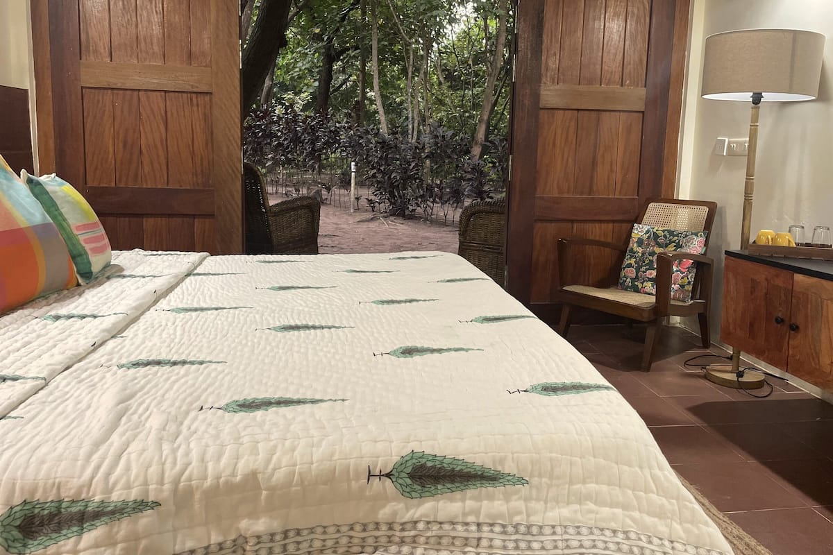 Una cama con colcha de estampado de hojas, una silla con cojín floral y una lámpara sobre una mesita de noche de madera.