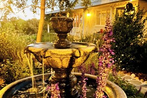 Une fontaine d’où jaillit de l’eau, entourée de verdure, avec un bâtiment en arrière-plan.
