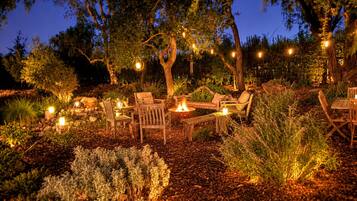 An outdoor seating area with wooden furniture and a fire pit, surrounded by trees and plants.