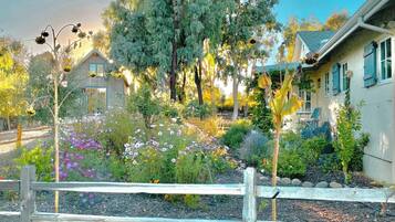 A residential garden with a wooden fence, a variety of flowering plants, and a house with a sloped roof and blue shutters.