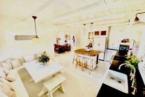 Una cocina moderna con una isla central, un comedor con mesa y banco blancos, y una sala de estar con sofá y mesa de centro.