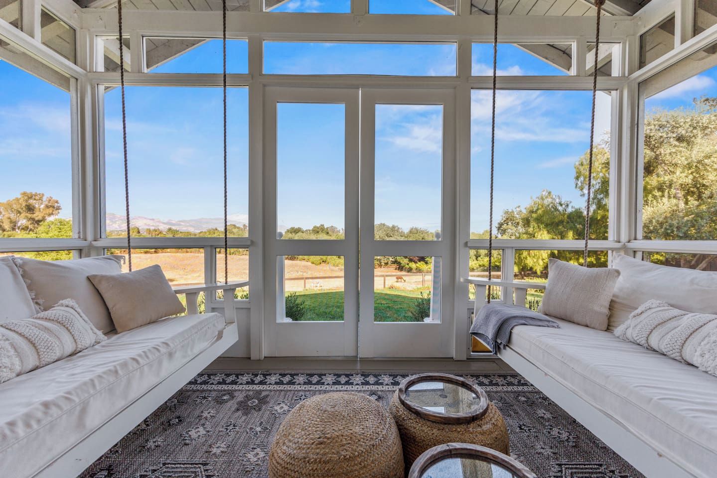 A room with two white sofas facing a large window, a woven ottoman, and a patterned rug.
