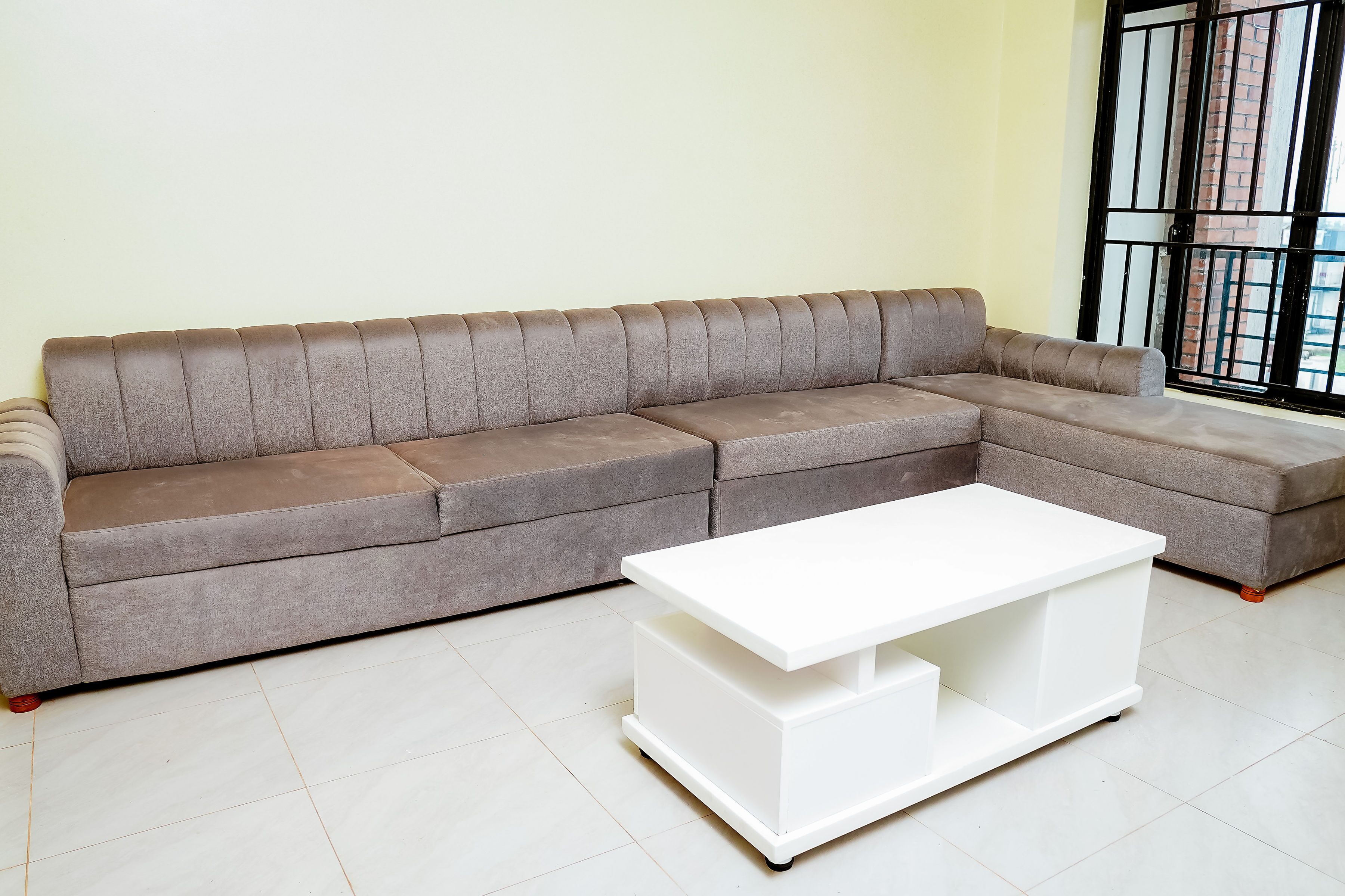 A grey sectional sofa with a white coffee table in a room with a tiled floor and a window with black bars.