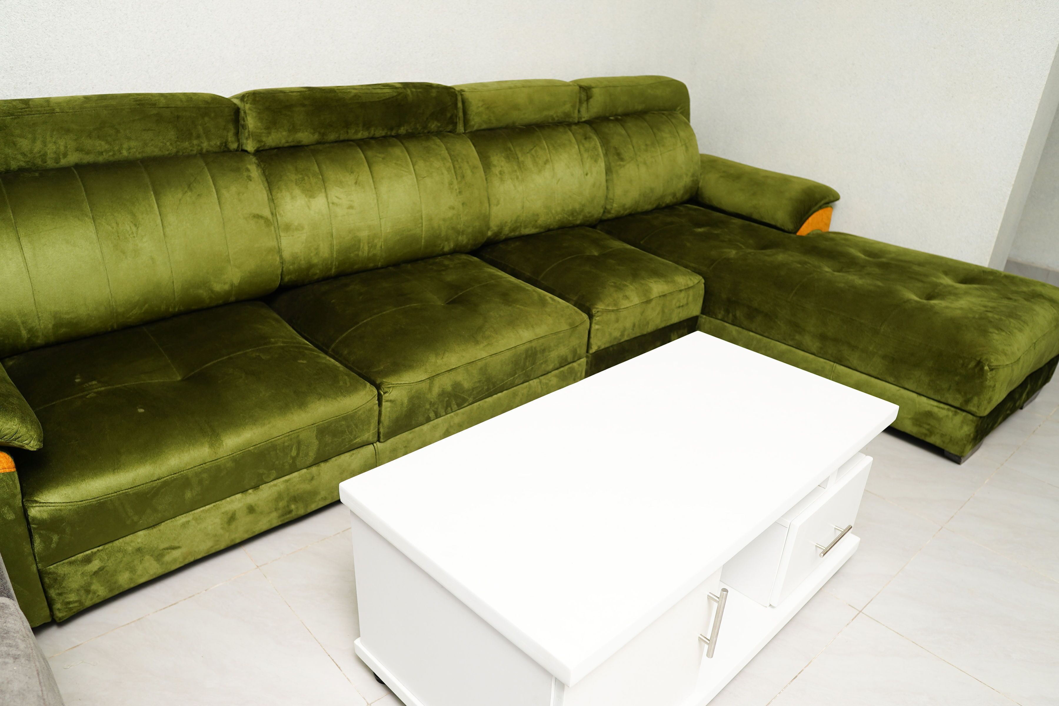 A green sectional sofa with a white coffee table in a room with white walls and tiled flooring.