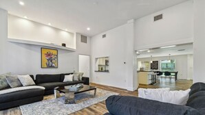 Ein modernes Wohnzimmer mit einem schwarzen Sofa, einem Couchtisch und einem Teppich. Im Hintergrund befindet sich ein Küchenbereich mit einem Esstisch und Stühlen.