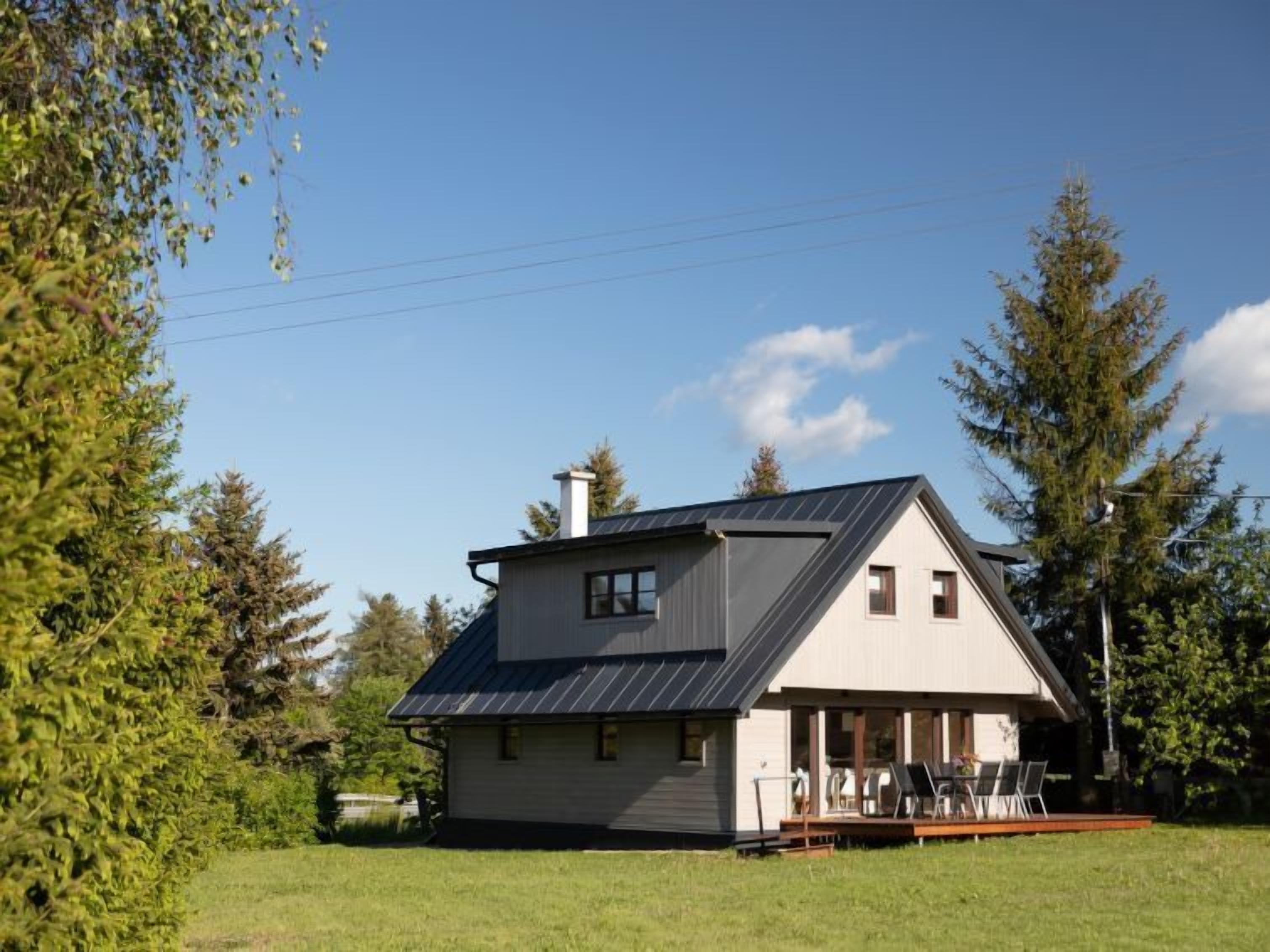 Ein modernes Haus mit Satteldach, einer überdachten Veranda mit Sitzgelegenheit und einem großen Garten, der von Bäumen umgeben ist.