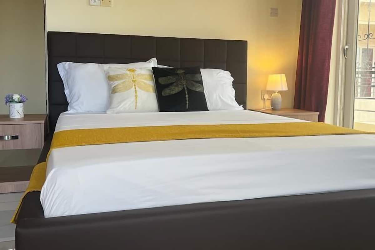 Una cama grande con cabecera negra, ropa de cama blanca y dorada, y un detalle en color amarillo.