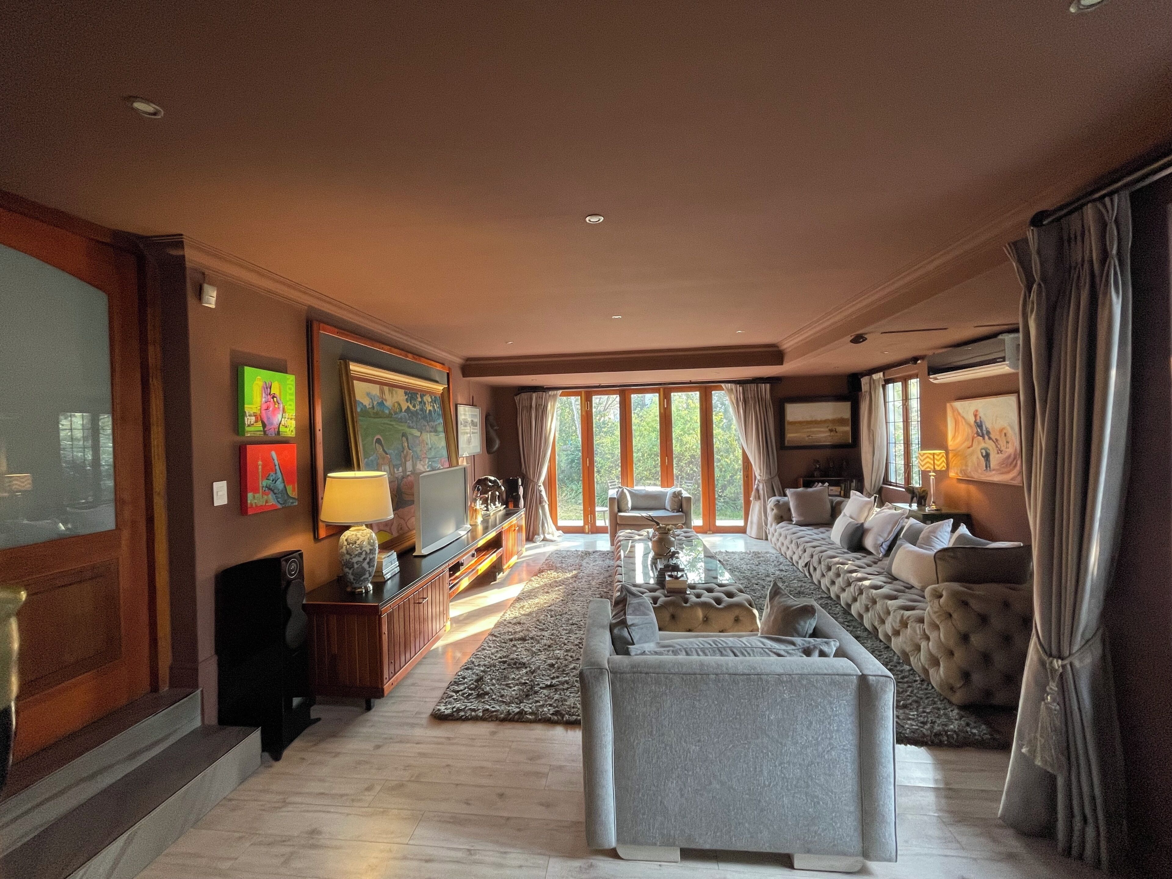 A spacious living room with a large sofa, a coffee table, and a television. There are large windows providing natural light and a view of the outdoors.