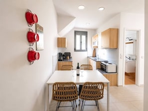 A modern kitchen with white cabinets, a dining table with wicker chairs, and a red kettle on the wall. - Urban Red (Rethymno)