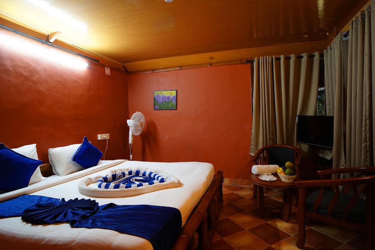 A hotel room with a bed, a chair, a table with fruits, a fan, and a painting on the wall.