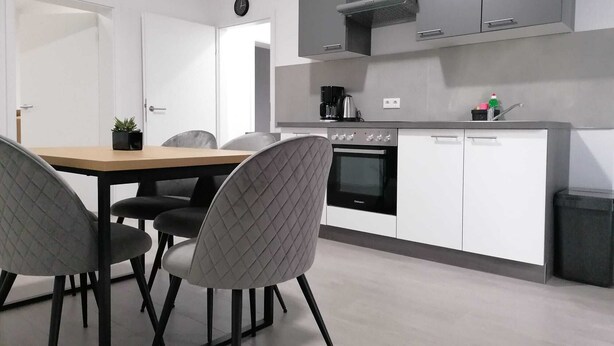 A modern kitchen with a built-in oven, a coffee maker, and a sink. There is a dining table with chairs and a clock on the wall. - estrella24 LIVING ROOMS Venice (Herne)