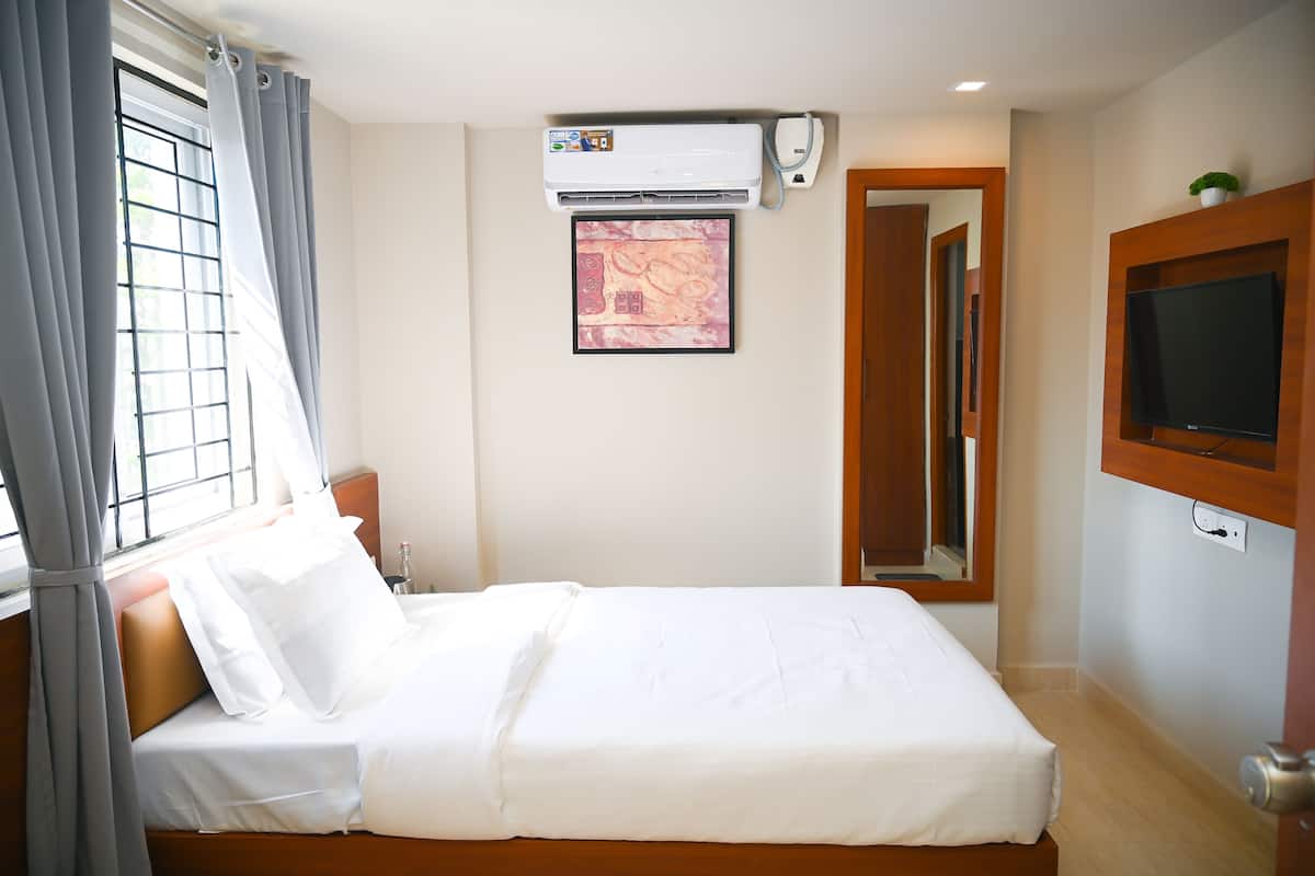 A neatly made bed with white linens, a wall-mounted air conditioner, a framed artwork, a television, and a wooden door.