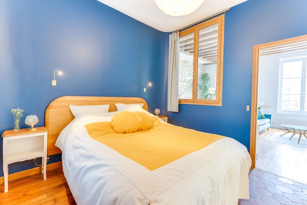 Een slaapkamer met een houten hoofdbord, een bed met wit en geel beddengoed, een nachtkastje met een lamp en een raam met gordijnen.